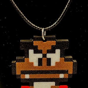 May include: A pixelated pendant necklace featuring a character from a classic video game. The pendant has a brown, black, white, and red color scheme. The necklace has a black cord and a silver-colored clasp.