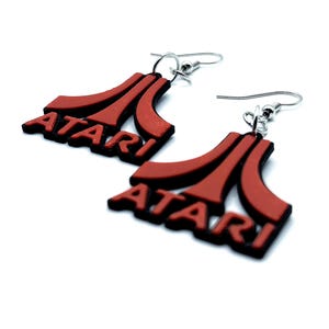 May include: A pair of red and black Atari logo earrings. The earrings are made of a layered material, with the Atari logo in red on top of a black base. The earrings have silver-colored fish hook ear wires.