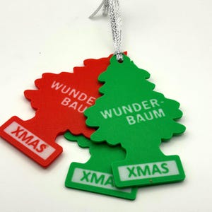 Op de afbeelding: Rode en groene kerstboomvormige ornamenten met de tekst "Wunder-Baum" en "XMAS" in het wit. De ornamenten zijn bevestigd aan een zilveren draad.
