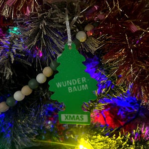 Op de afbeelding: Een groene kerstboomvormige ornament met de woorden "WUNDER-BAUM" en "XMAS" in het wit. De ornament hangt aan een zilveren draad en wordt getoond op een versierde kerstboom met kleurrijke lichten en slingers.