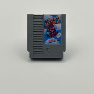 May include: A gray Super Mario Bros. 2 video game cartridge. The label features Mario in a red outfit with a blue background and the game title. The cartridge is a classic Nintendo Entertainment System (NES) game.