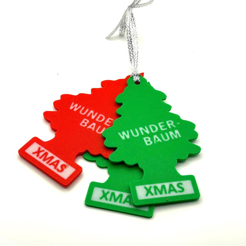Op de afbeelding: Rode en groene kerstboomvormige luchtverfrissers met de woorden "Wunder-Baum" en "XMAS" erop gedrukt. Ze hangen aan een zilveren lint. De achtergrond is wit.