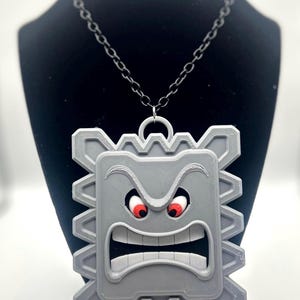 May include: A gray Thwomp pendant necklace. The pendant features a rectangular shape with a menacing face, red eyes, and a wide open mouth. The necklace has a black chain.