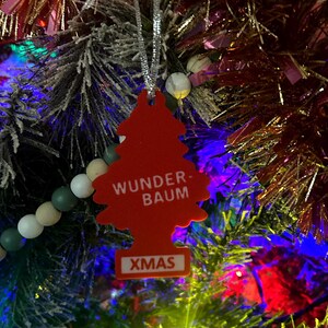 Op de afbeelding: Een rode, kerstboomvormige luchtverfrisser met de woorden "WUNDER-BAUM" en "XMAS" erop gedrukt, hangend aan een versierde kerstboom. De boom heeft kleurrijke lichten en ornamenten.