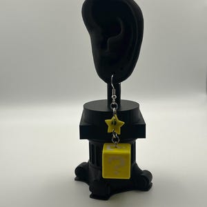 以下が含まれることがあります： 黄色い星とクエスチョンマークの付いた黄色の立方体が付いたシングルイヤリング。イヤリングはシルバーのフックから吊り下げられています。イヤリングは黒い耳の形をしたスタンドに展示されています。