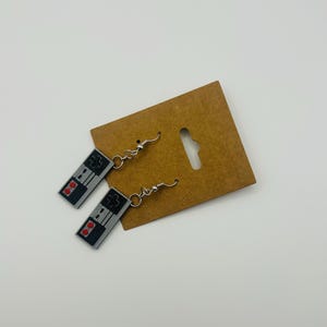 May include: A pair of silver-toned earrings featuring a retro video game controller design. The controllers are gray with black and red details, hanging from silver fish hook ear wires. The earrings are displayed on a brown cardboard backing.