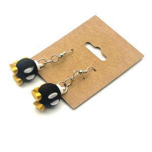 Puede incluir: Un par de pendientes negros y blancos con forma de bomba y detalles dorados. Los pendientes están sujetos a ganchos para la oreja de color plateado. Los pendientes se muestran en una tarjeta de exhibición de pendientes de cartón marrón.