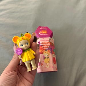 May include: A small, collectible figurine from the Sonny Angel Pumpkin Patch Series. The figure is dressed in a yellow pumpkin costume and holds a purple lollipop. The packaging is visible, featuring the series name and illustrations.