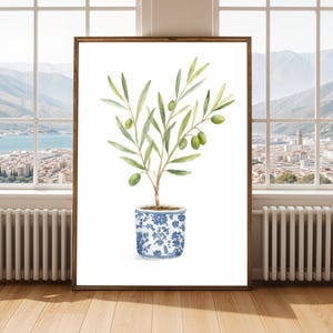May include: Watercolour painting of an olive tree in a blue and white floral pot. The artwork is framed in a brown wooden frame. The olive tree has green leaves and olives. The background shows a city and mountains.