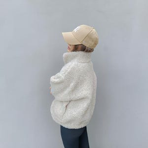 Peut inclure: Une casquette de baseball beige avec un logo NY blanc et une veste en polaire crème à col montant et manches longues, dotée d'une surface texturée et duveteuse. La veste présente une fermeture éclair frontale et une coupe ample et décontractée.