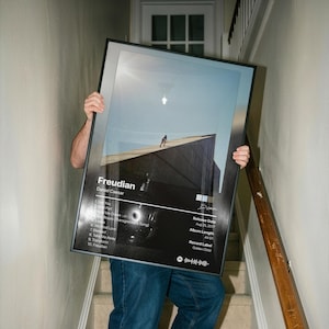 May include: A framed rectangular poster featuring the album cover for Freudian by Daniel Caesar. The design includes a photograph of a person on a roof, a tracklist of ten songs, release date August 25, 2017, album length 44:51, and record label Golden Child.