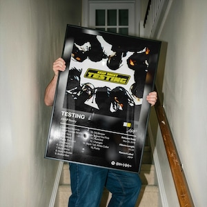 Puede incluir: Un póster enmarcado con la portada del álbum "Testing" de ASAP Rocky. El póster tiene un marco negro y muestra una imagen en blanco y negro con la palabra "TESTING" en amarillo y negro. La lista de canciones del álbum también es visible.