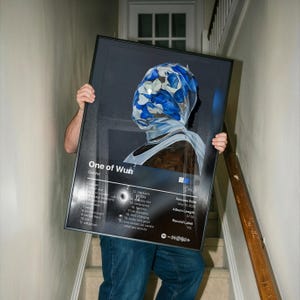 May include: A framed poster featuring the album art for "One of Wun" by Gunna. The artwork depicts a person wearing a blue and white patterned head covering. The poster includes the album's tracklist, release date, and record label.