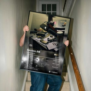 May include: A framed poster featuring the album cover art for Kendrick Lamar's Section.80. The design includes a tracklist, release date, and album length on a black background with a photograph of various items including a gold chain.