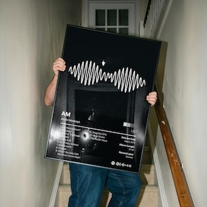 May include: A rectangular framed poster featuring the Arctic Monkeys AM album artwork. The black background displays a white soundwave graphic, the text AM, tracklist, release date Sep 9, 2013, album length 41:47, record label Domino, and a Spotify code.