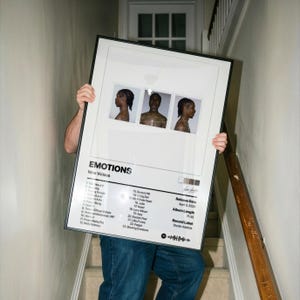 May include: A framed poster titled "EMOTIONS" by Nine Vicious. The poster features three small photos of a person with wet hair. The poster includes a tracklist, release date, album length, and record label. The poster is held by a person on a staircase.