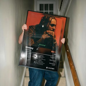 Può includere: Un poster rettangolare incorniciato che presenta la copertina dell'album Stoney di Post Malone. L'opera mostra un ritratto dell'artista su uno sfondo arancione, con una tracklist, la data di uscita e la durata dell'album in testo bianco.