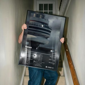 May include: A framed poster featuring the album "Views" by Drake. The poster has a black frame and displays the album cover art, a grayscale image of a building. The tracklist and release details are also visible.