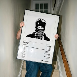 Op de afbeelding: Een zwart-wit ingelijste poster met een gestileerde afbeelding van een persoon met een hoed en gezichtsbedekking. De poster bevat de titel "Rebel" en de artiestennaam EsDeeKid, samen met een tracklist en release-informatie.