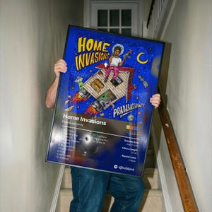 May include: A framed poster titled "Home Invasions" by Pradabagshawty. The artwork features a house-shaped spaceship with a person in a spacesuit. The poster has a blue background with the artist's name and song titles.