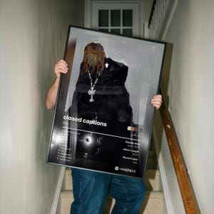May include: A framed poster with the text "closed captions" and the artist's name "Che". The artwork features a person in black clothing and a silver cross necklace. The poster also includes a tracklist, release date, and album length.