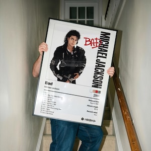 May include: Framed poster featuring Michael Jackson's "Bad" album cover. The image shows Jackson in a black jacket. The poster includes the album title, tracklist, release date, and record label information. The poster is held in front of a person on a staircase.