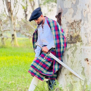 Può includere: Una persona in abito tradizionale scozzese, con un kilt a quadri, uno scialle abbinato, una camicia bianca, un basco nero e stivali neri. L'outfit è completato da una lunga spada. Il kilt presenta un motivo a quadri rossi, verdi e blu.