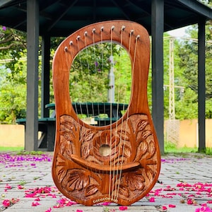 Puede incluir: Una lira de madera hecha a mano con intrincadas tallas florales. El instrumento presenta un marco curvo, cuerdas y clavijas de afinación. La lira es de color marrón cálido y descansa sobre una superficie de ladrillo con pétalos de flores rosas dispersos.