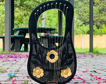 Op de afbeelding: Een zwarte lier met goudkleurige bloemaccenten en snaren. Het instrument is versierd met ingewikkelde bloempatronen en staat tegen een vage buitenachtergrond met een prieel en roze bloemblaadjes.