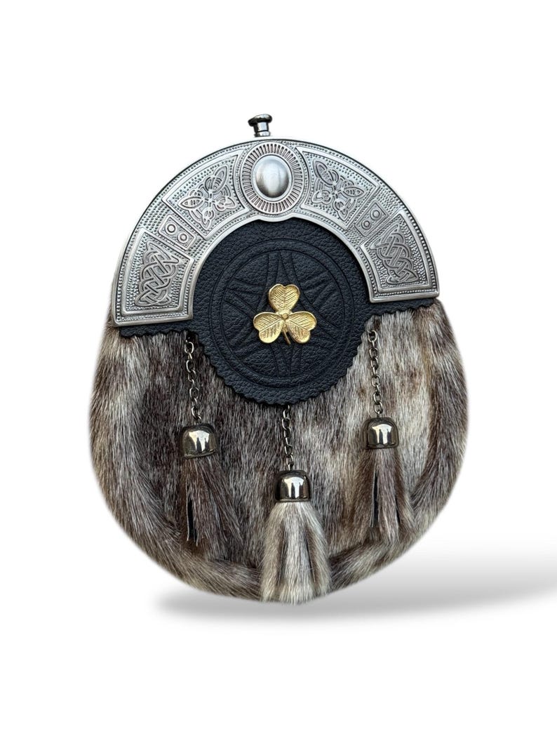 K&ouml;nnte beinhalten: Eine traditionelle schottische Sporran mit silbernem Cantle und Fellk&ouml;rper. Der Cantle zeigt keltische Knotenmuster und einen zentralen Stein. Die schwarze Lederklappe hat ein goldenes Kleeblatt-Emblem. Die Sporran hat drei Quasten mit silbernen Kappen und Ketten.