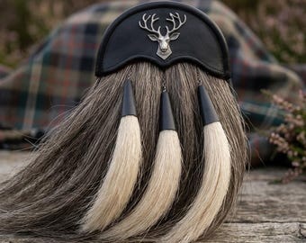 Könnte beinhalten: Ein schwarzer Leder-Sporran mit silbernem Hirsch-Emblem, grauem Fellkörper und drei weißen Quasten mit schwarzen Spitzen. Der Hintergrund zeigt einen Tartan-Stoff und eine Holzoberfläche.