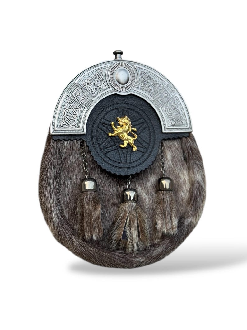 K&ouml;nnte beinhalten: Eine traditionelle schottische Sporran mit einem silbernen Cantle mit keltischen Knoten und einem zentralen Stein. Die schwarze Lederklappe hat ein goldenes L&ouml;wenemblem. Der K&ouml;rper besteht aus braunem Fell mit drei passenden Quasten.