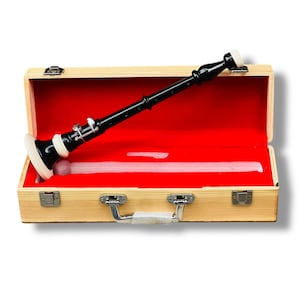 Könnte beinhalten: Eine schwarz-weiße Oboe liegt in einem Holzkoffer mit rotem Samtinnenfutter. Die Oboe hat silberne Klappen und ein weißes Mundstück. Der Koffer ist rechteckig mit einem silbernen Griff und Verschlüssen.
