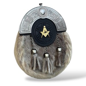 K&ouml;nnte beinhalten: Ein traditioneller schottischer Sporran mit einem silbernen Cantle mit keltischen Knoten und einem zentralen Stein. Der schwarze Lederk&ouml;rper hat ein goldenes Freimaurersymbol. Der Sporran hat einen Pelzk&ouml;rper und drei silberne Quastenanh&auml;nger.