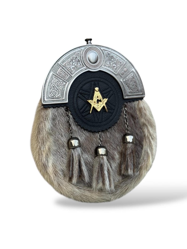 K&ouml;nnte beinhalten: Ein traditioneller schottischer Sporran mit einem silbernen Cantle mit keltischen Knoten und einem zentralen Stein. Der schwarze Lederk&ouml;rper hat ein goldenes Freimaurersymbol. Der Sporran hat einen Pelzk&ouml;rper und drei silberne Quastenanh&auml;nger.