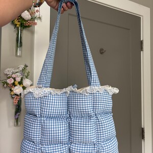 以下が含まれることがあります： 青と白のギンガムチェック柄のキルティングトートバッグ。柔らかくふっくらとした四角いステッチ模様が特徴です。バッグの上端には繊細な白いレースのトリムがあしらわれており、持ち運び用の共布のショルダーストラップが2本付いています。