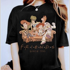May include: Black t-shirt featuring a graphic of Toy Story characters on a couch. The design includes Woody, Buzz Lightyear, and other characters. The text "F.R.I.E.N.D.S" and "SINCE 1995" are printed below the graphic.