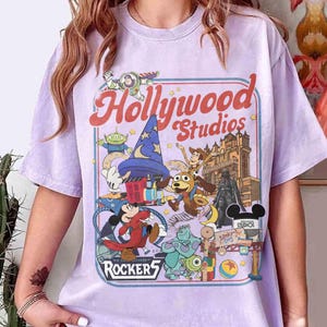 Może przedstawiać: Jasnofioletowa koszulka z kolorową grafiką z Disney's Hollywood Studios. Projekt zawiera postacie takie jak Myszka Miki, Buzz Astral i Darth Vader, z widocznymi napisami "Hollywood Studios" i "Rockers 5".