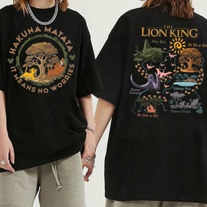 May include: Black t-shirts featuring graphics from The Lion King. One shirt has a circular design with Simba, Timon, and Pumbaa, with the text "Hakuna Matata". The other shirt has a map-like design with various scenes and the text "The Lion King".