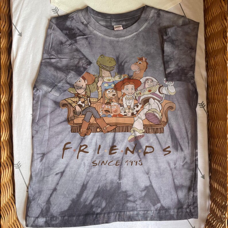May include: Gray tie-dye t-shirt featuring a graphic of characters from the animated film "Toy Story" sitting on a couch. The word "FRIENDS" is printed in brown letters, with "SINCE 1995" below.