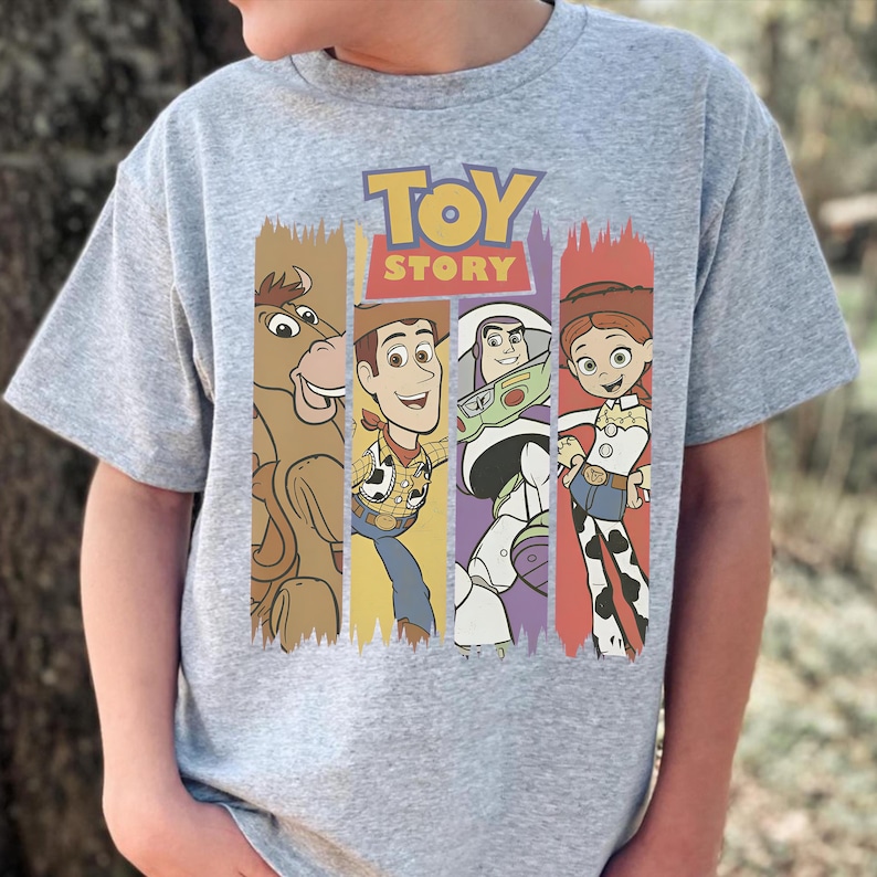 May include: A gray t-shirt featuring the words "TOY STORY" above cartoon illustrations of characters. The characters include a horse, Woody, Buzz Lightyear, and Jessie, each within a vertical colored stripe.