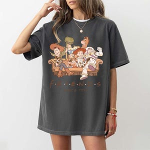 May include: Dark gray t-shirt featuring a graphic of Toy Story characters on a brown couch. The text "F.R.I.E.N.D.S. Since 1995" is printed below the image. The shirt has short sleeves and a relaxed fit.