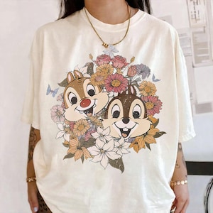 May include: Cream-colored t-shirt featuring a graphic of Chip and Dale surrounded by a floral arrangement in shades of pink, orange, and white. The design includes butterflies and is printed on a short-sleeved tee.