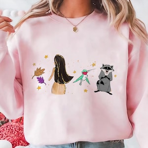 May include: A light pink sweatshirt featuring a cartoon illustration. The design includes a woman with long black hair, a hummingbird, a raccoon, leaves, and stars. The sweatshirt is a crew neck style.