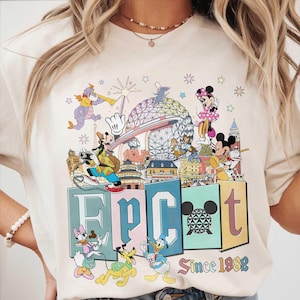 May include: Cream-colored t-shirt featuring a colorful graphic of Disney characters and landmarks. The word "EPCOT" is displayed in large letters, with "Since 1982" below. The design includes Mickey Mouse, Minnie Mouse, and Donald Duck.