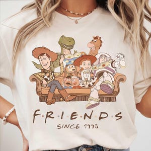 May include: Cream-colored t-shirt featuring a graphic of Toy Story characters sitting on a couch. The design includes Woody, Buzz Lightyear, and other characters. The text "FRIENDS SINCE 1995" is printed below the graphic.