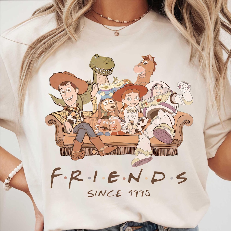 May include: Cream-colored t-shirt featuring a graphic of Toy Story characters sitting on a couch. The design includes Woody, Buzz Lightyear, and other characters. The text "FRIENDS SINCE 1995" is printed below the graphic.