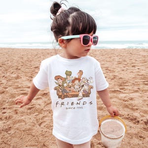 May include: A white t-shirt featuring a graphic of Toy Story characters sitting on a couch, with the word "FRIENDS" and "SINCE 1995" printed below. The child is wearing pink sunglasses and standing on a sandy beach.