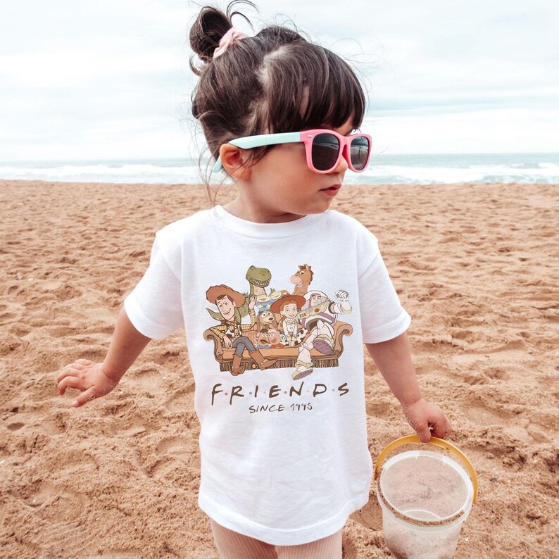 May include: A white t-shirt featuring a graphic of Toy Story characters sitting on a couch, with the word "FRIENDS" and "SINCE 1995" printed below. The child is wearing pink sunglasses and standing on a sandy beach.