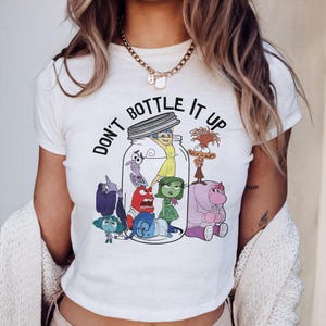 Peut inclure: T-shirt blanc avec un motif des personnages du film *Vice-Versa* dans un bocal et le texte "DON'T BOTTLE IT UP" en noir. Le t-shirt a un col rond et des manches courtes.